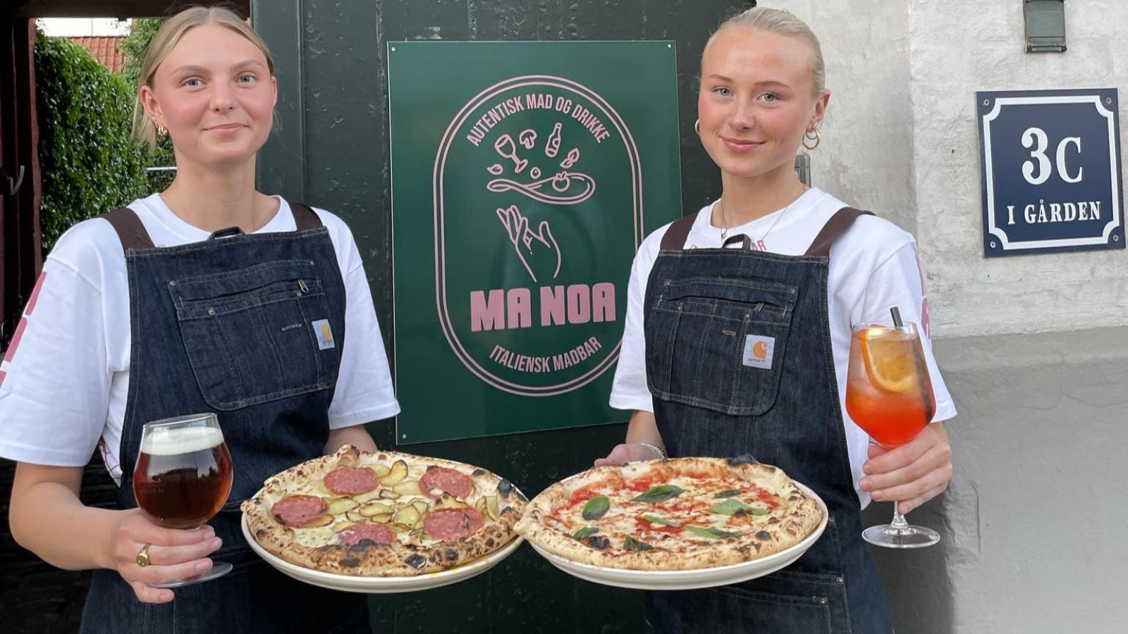 Zwei Kellner halten jeweils eine Pizza, ein Bier und ein Getränk.