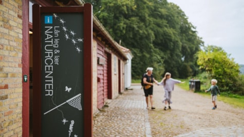 Familie går udenfor ved Naturcenter Trelde Næs. I forgrunden ses naturcentrets skilt.