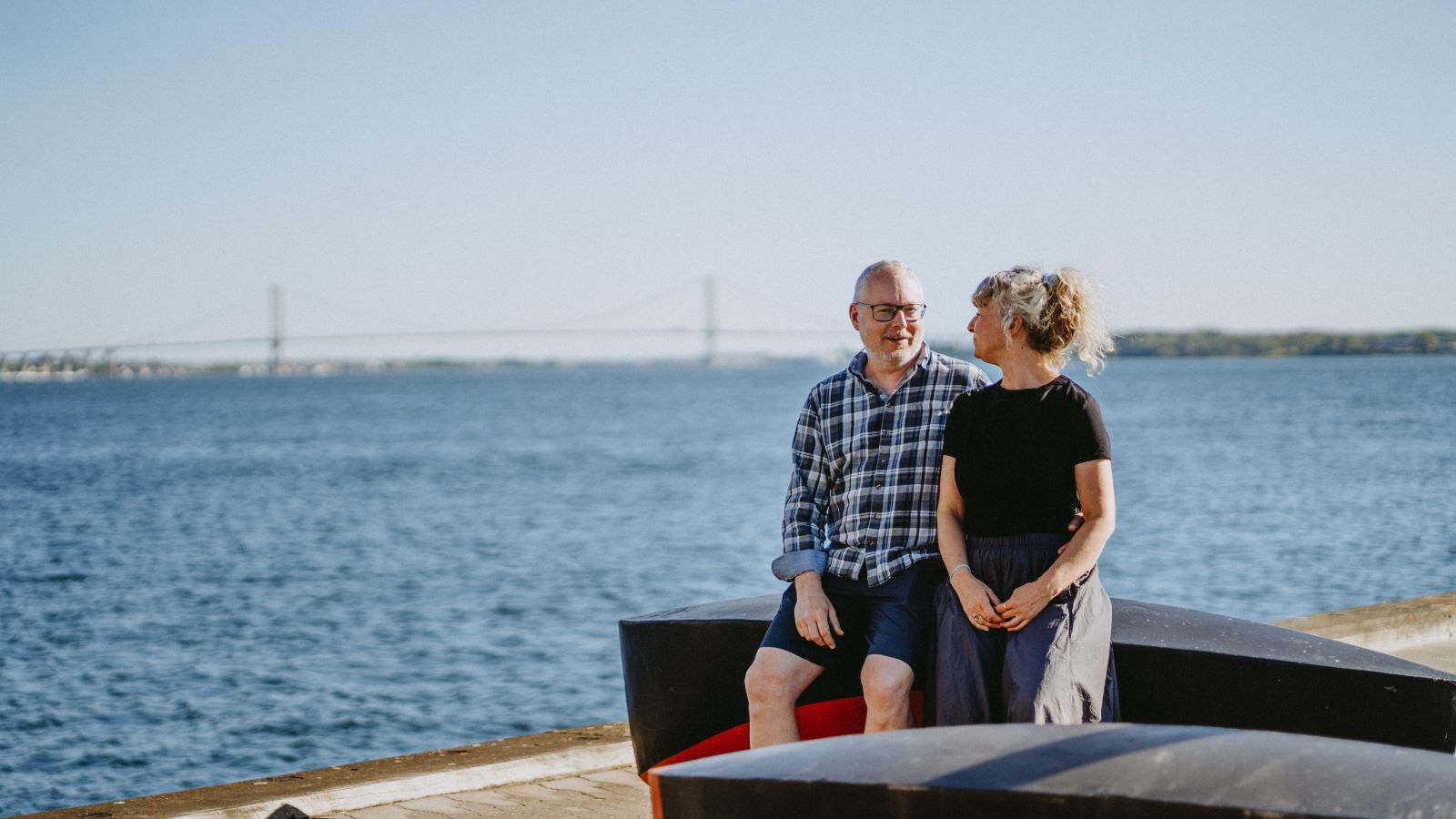 A couple sits on an artwork with a view of the Little Belt. In the background, the New Little Belt Bridge can be seen.