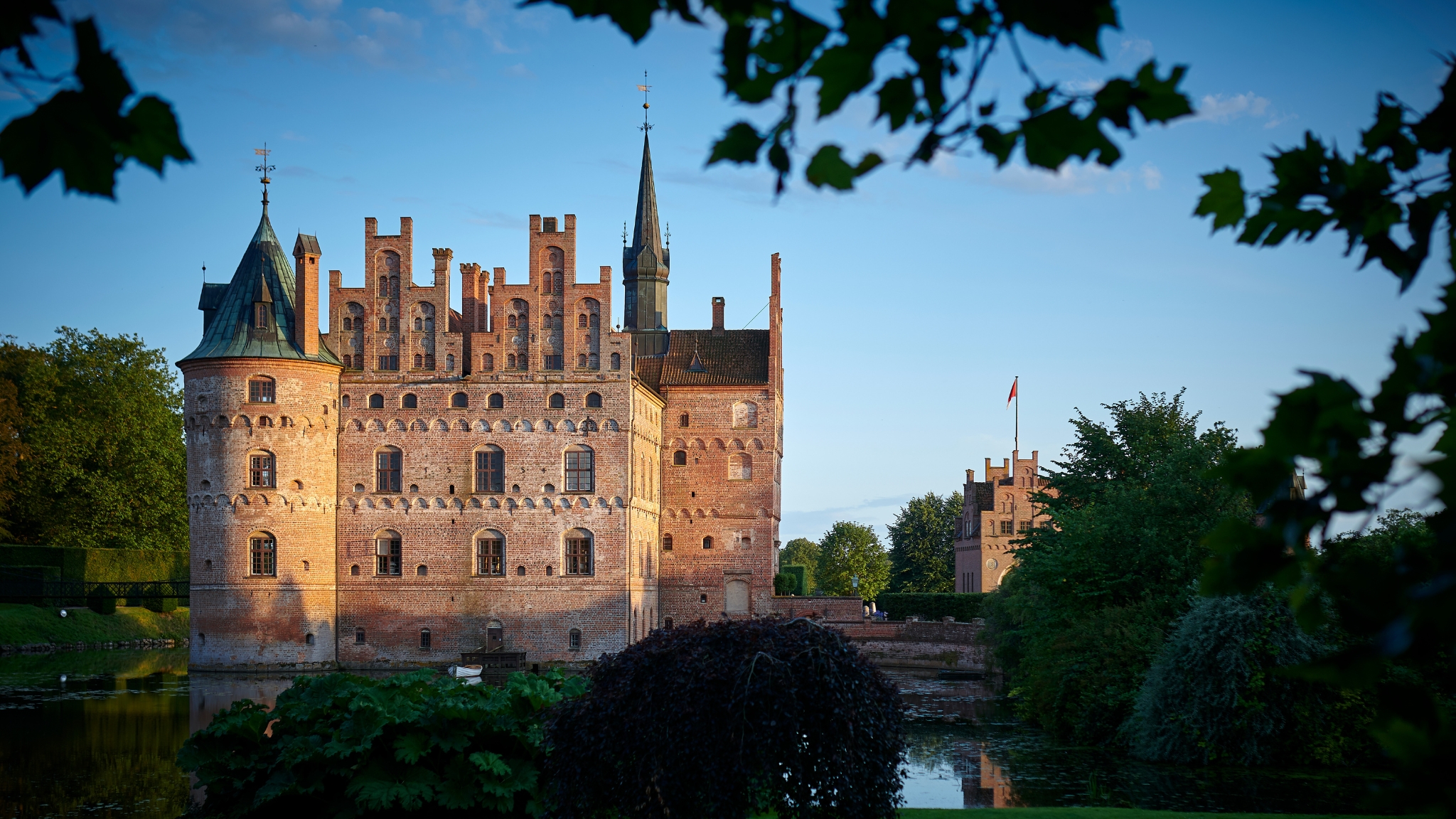 Egeskov Castle in the evening sun, seen from the moat, surrounded by green trees and reflections on the water.