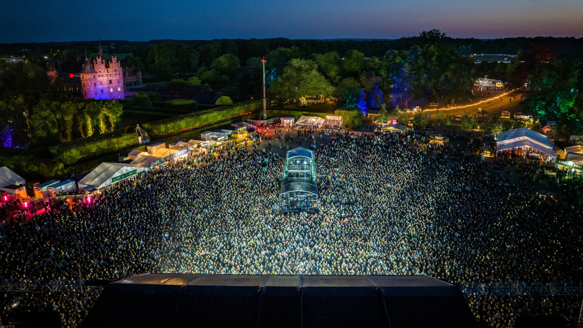 The Heartland Festival at Egeskov Castle, seen from above, with a large crowd, the stage and lights in the twilight.