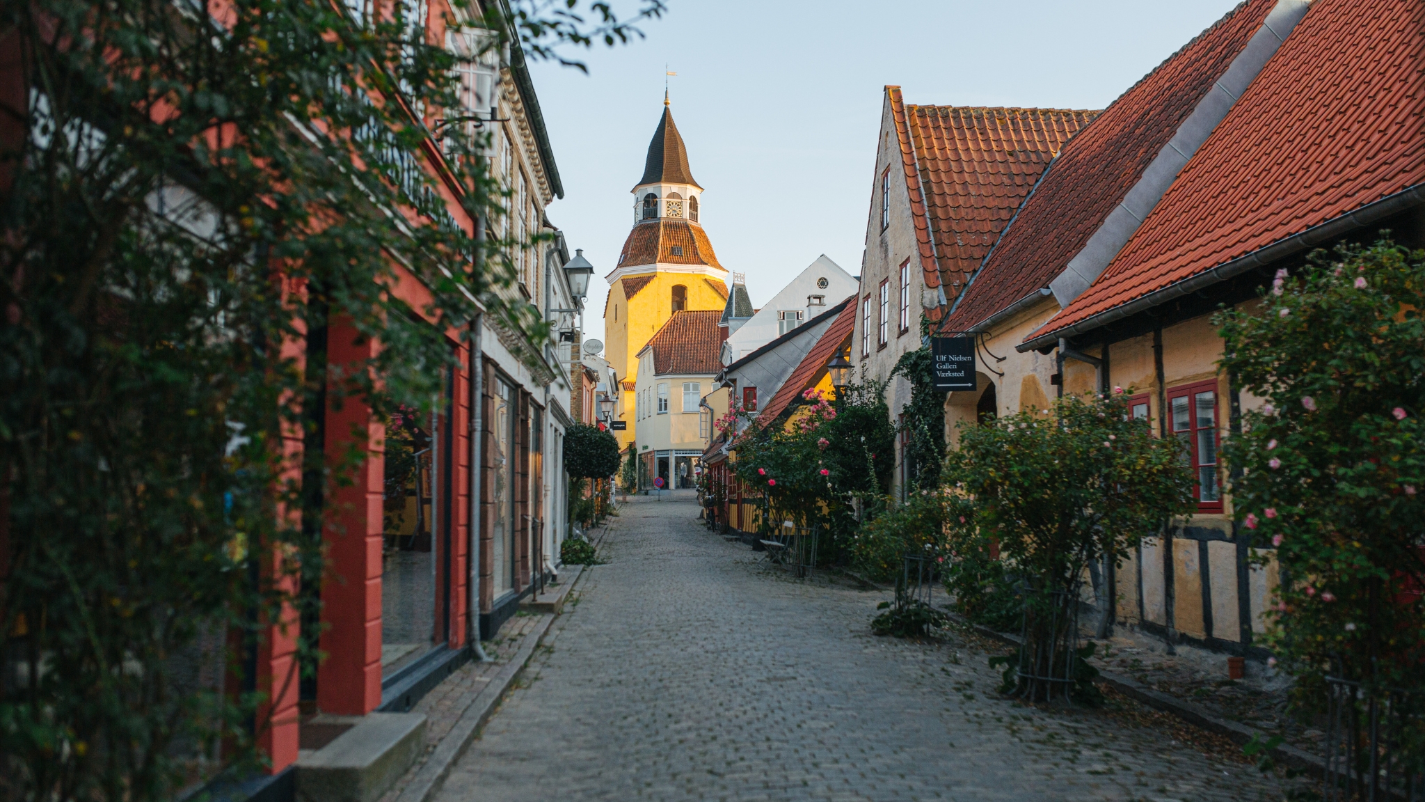 Kopfsteinpflastergasse mit alten Häusern, Stockrosen und dem Glockenturm von Faaborg im Hintergrund.