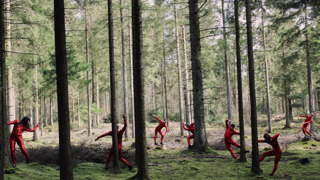 Otte dansere danser i en skov, ved hver sin træstamme. Alle har rødt tøj på