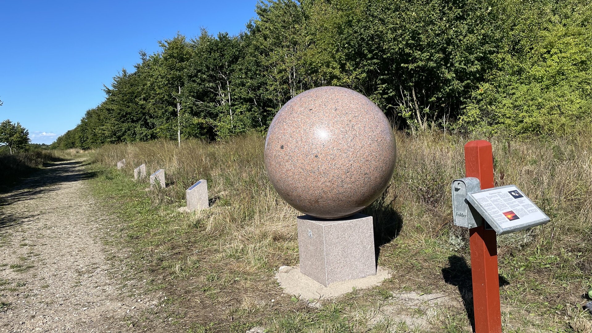 Große rötliche Granitkugel auf einem Sockel an einem Kiesweg, daneben kleinere Granitsteine in einer Reihe und eine Infotafel an einem roten Pfosten.