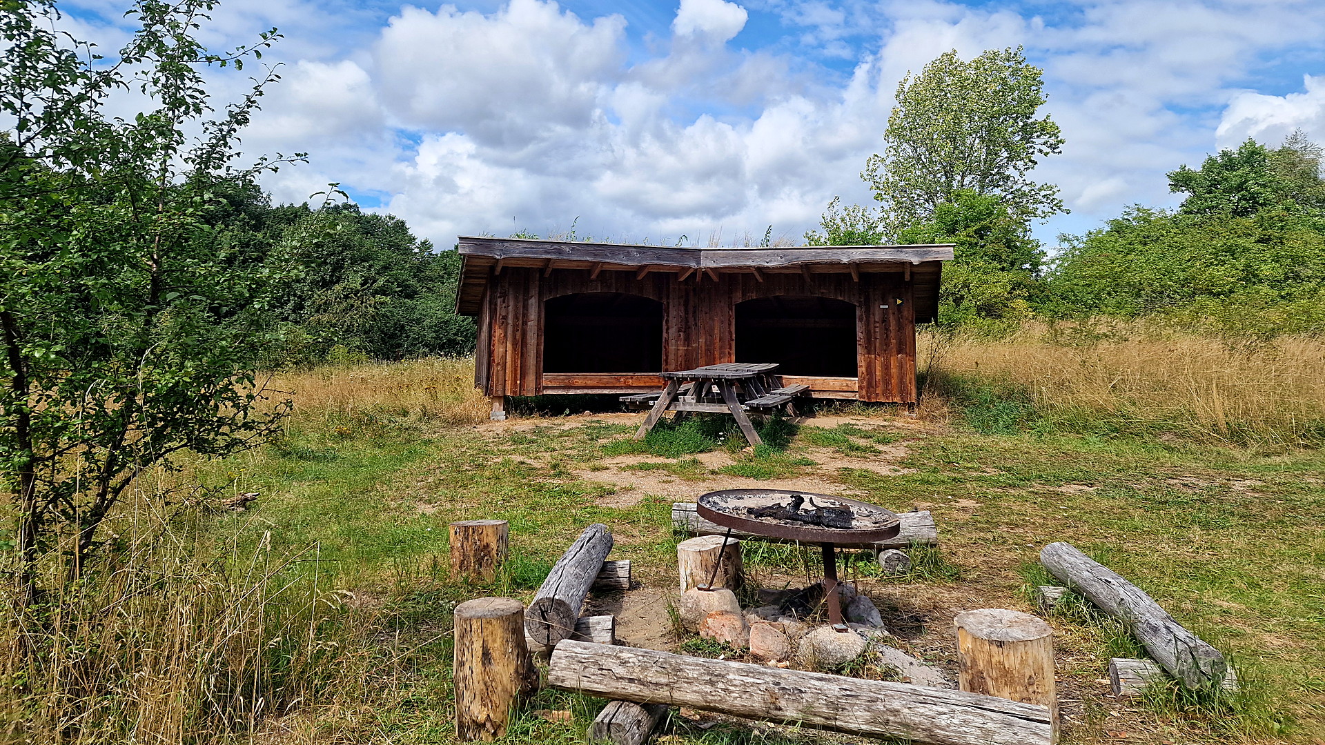 Fire pit with grill grate and log seating in front of the shelter in the meadow at Skellingsted outdoor area near Mørkøv.