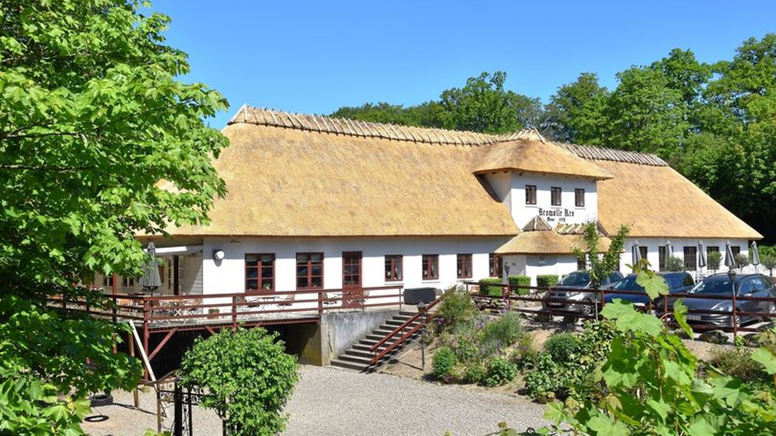 Exterior view of Bromølle Kro – whitewashed inn with thatched roof, wooden terrace and courtyard surrounded by forest.