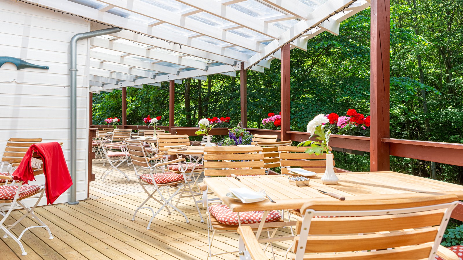 Covered terrace at Bromølle Kro with wooden tables, geraniums and a view of the forest.