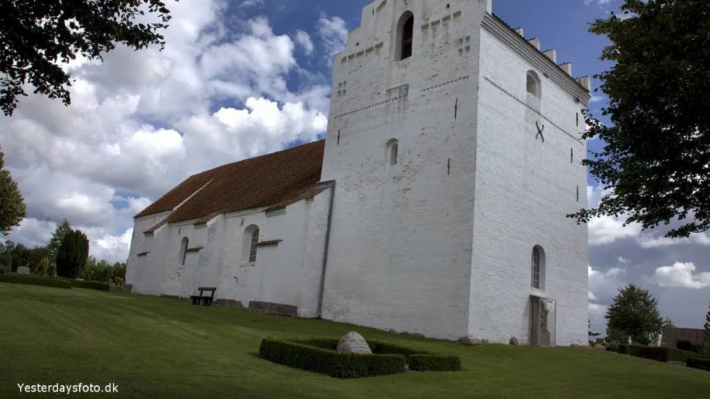The white-washed Drigstrup Church against a backdrop of dark clouds, with a well-maintained cemetery and tall trees.