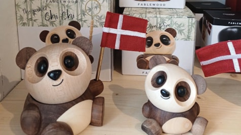 To pandaer i træ sidder på et bord. Pandaerne har to forskellige størrelser, den største har et flag i hånden