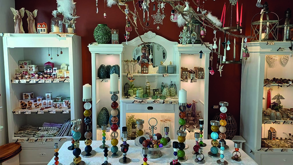 Table full of candlesticks, the old-fashioned shelves in the background are filled with knick-knacks and Christmas decorations for the home. From the ceiling hang Christmas ornaments and baubles.