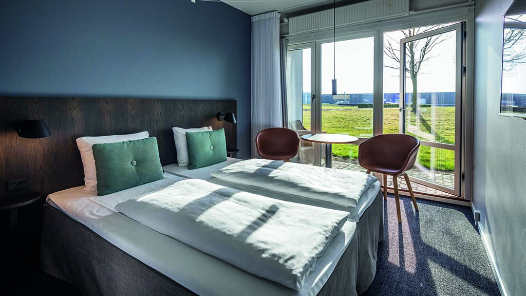 Room with a large window overlooking a spacious green lawn. Sunlight streams through the window into the room. There is a double bed and two chairs in the room.
