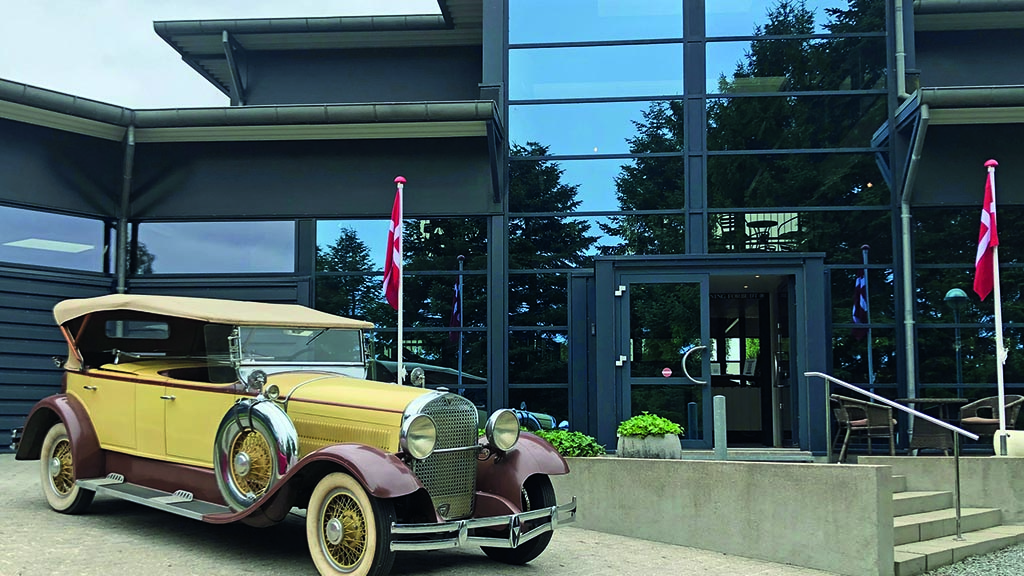 Vor Strib Automobile Museum steht ein gelber Oldtimer aus den 1920er- oder 1930er-Jahren. Das Museum verfügt über großen fenstern und zwei Dannebros-Flaggen am Eingang.