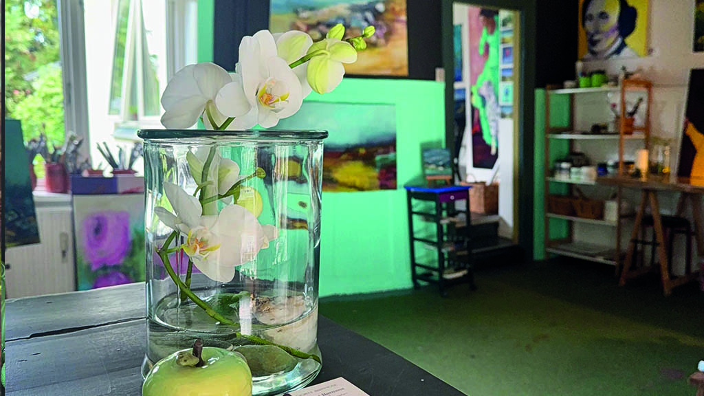 i forgrunden er et bord med en glas vase med en orkide i. I baggrunden ses Galleriet med hylder og billeder på væggene