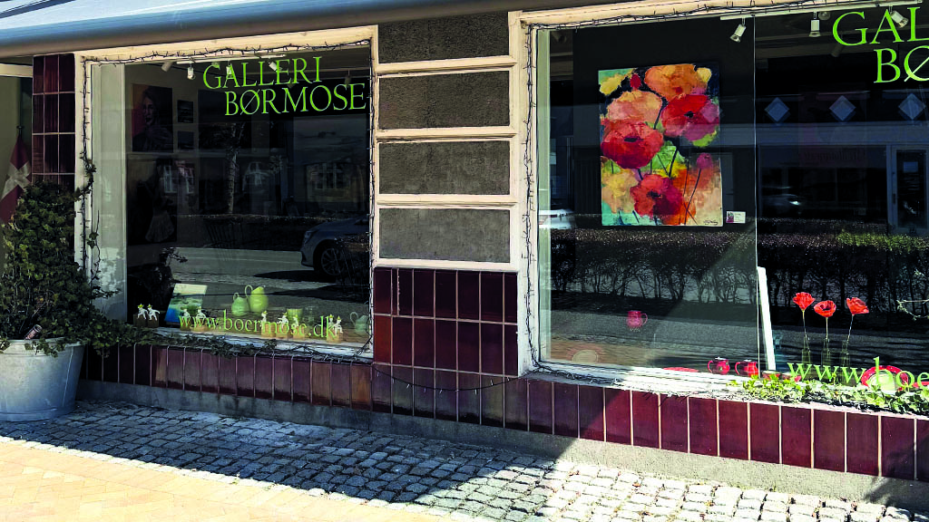 Galleri Børmoses facade på en solskinsdag. Der er to store glas vinduer og man kan se et maleri af nogle blomster i det ene