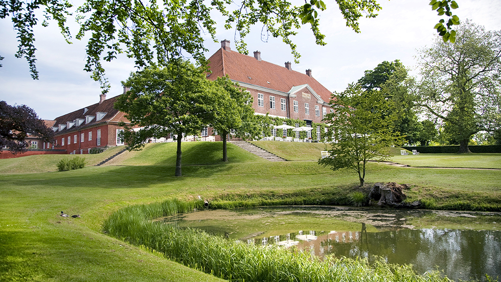 Hindsgavl Castle in the background, a large lake in the foreground. Lush and green. Hindsgavl Castle is built of red brick with a red tile roof.
