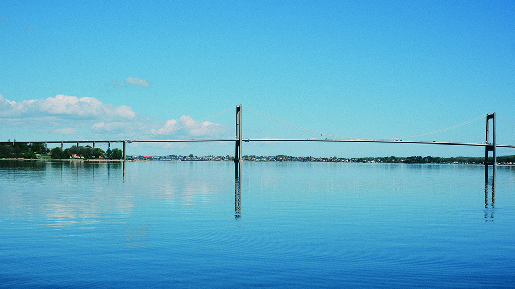 Den nye Lillebæltsbro spejler sig i vandet ved Lillebælt. Der er en sommerdag vandet er stille og himlen er blå med få skyer
