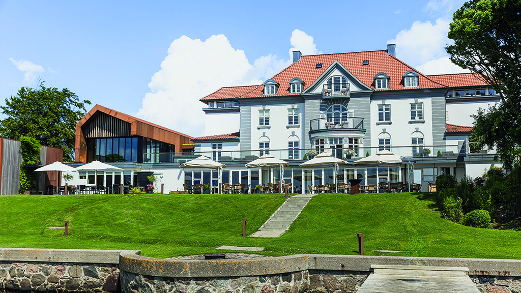 På billedet ses hotel Sixtus. Billedet er taget fra vandet og man kigger op mod hotellet. Foran bygningen er en terrasse med parasoller og en stor grøn græsplæne