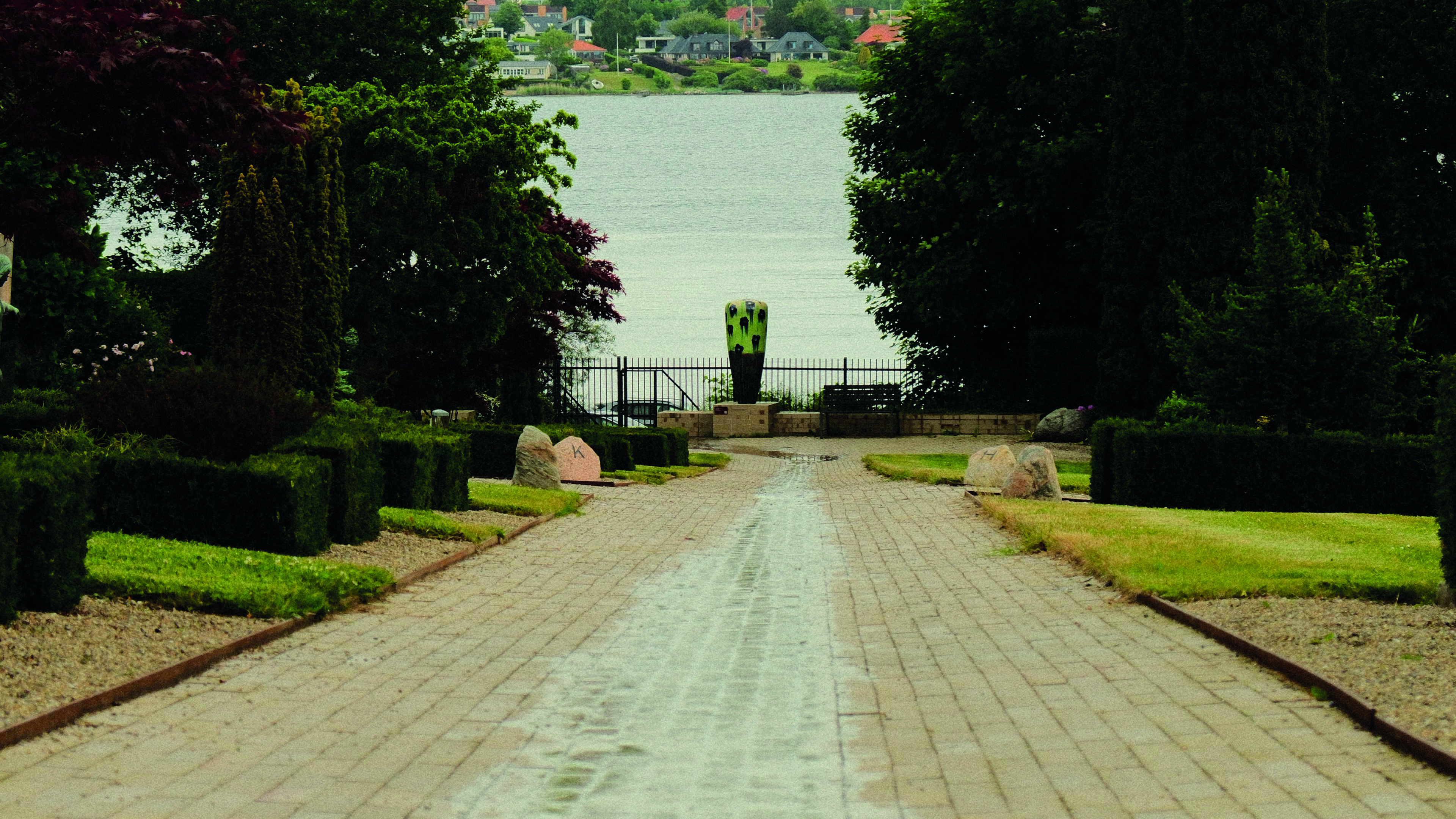 Regenwasser fließt mittig über eine Trittstufe auf einem Friedhof. Am Ende steht eine große Keramikvase, und im Hintergrund ist Lillebælt zu sehen.
