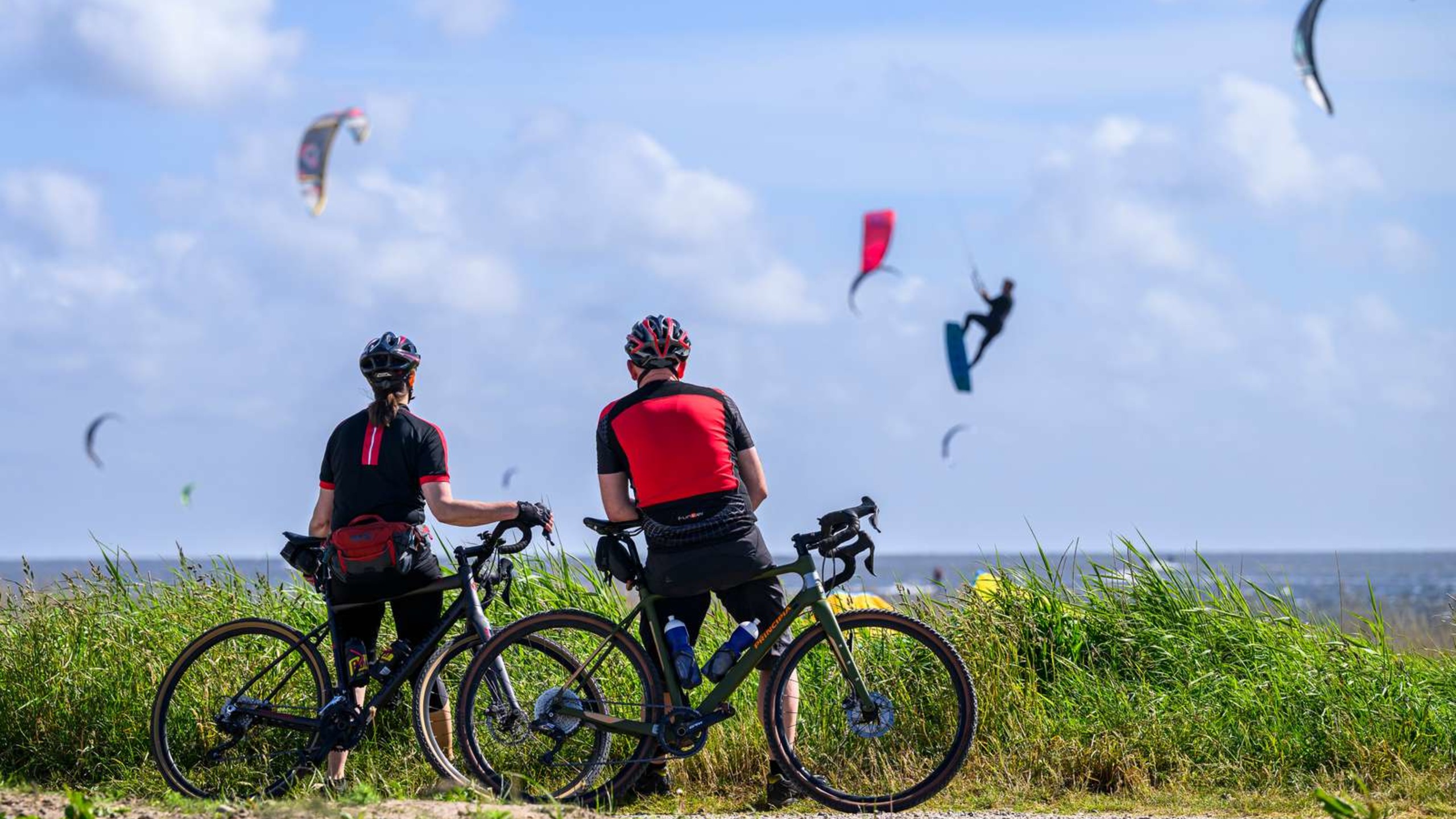 Two cyclists on a nature trail by the fjord, looking out towards the kitesurfers on the fjord.