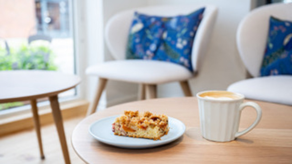 På et rundt bord står der en tallerken med kage og en kop kaffe i hyggelige omgivelser med stole rundt om.