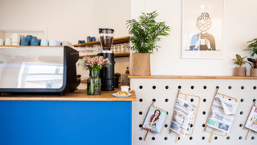En blå disk med kaffemaskine, blomster og kopper på pryder venstre side. Til højre for den ligger der nogle aviser og et billede, der skaber en afslappet stemning.