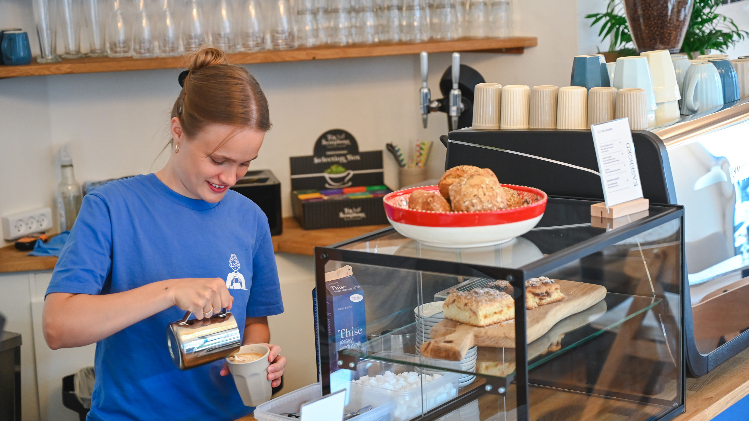A café counter with cups and glasses, behind which a member of staff is making coffee.