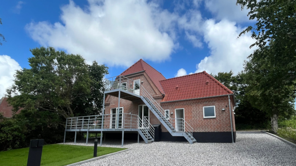 Bagsiden af et rødt murstenshus med trappeopgang og altan, der fører ud til flere værelser. Der ligger også en indkørsel og en lille have.