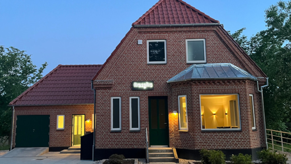 Et rødt murstenshus, hvorfra der stråler et hyggeligt lys ud af vinduerne. Med hvid bogstaver står der "fireogfirs" på huset.