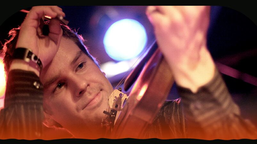 Foto af en musiker med et strygeinstrument i hånden og scenelys i baggrunden.