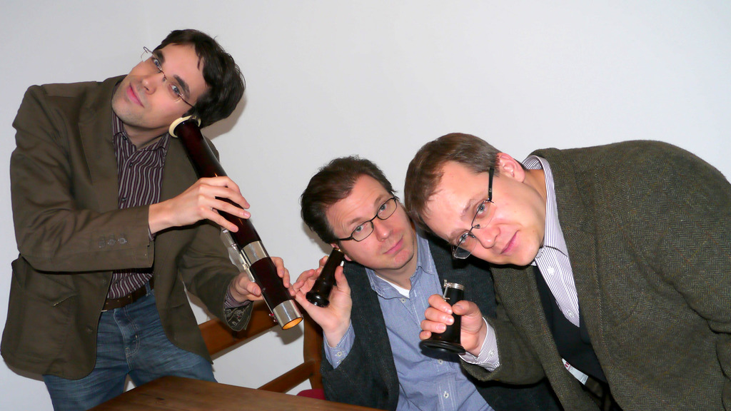 Three musicians are each holding a wind instrument and posing close together in front of a light-coloured background. The instruments are held up to their ears like telephone receivers.