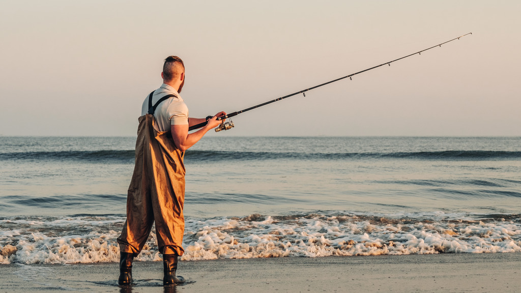 A man is standing in the surf by the sea, fishing.