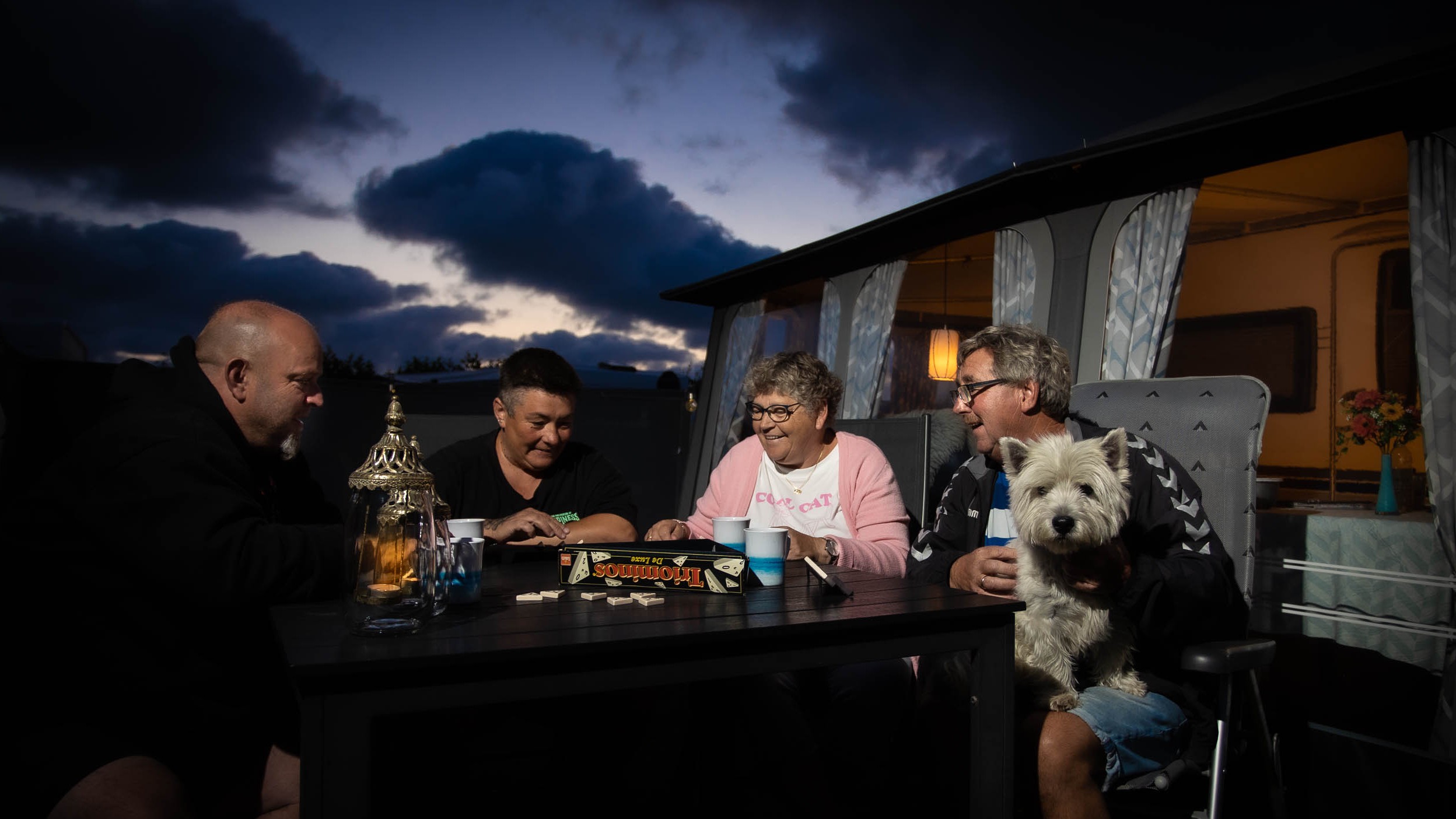 Two generations and a dog, absorbed in a game of Triominos at the camping table in the evening light.