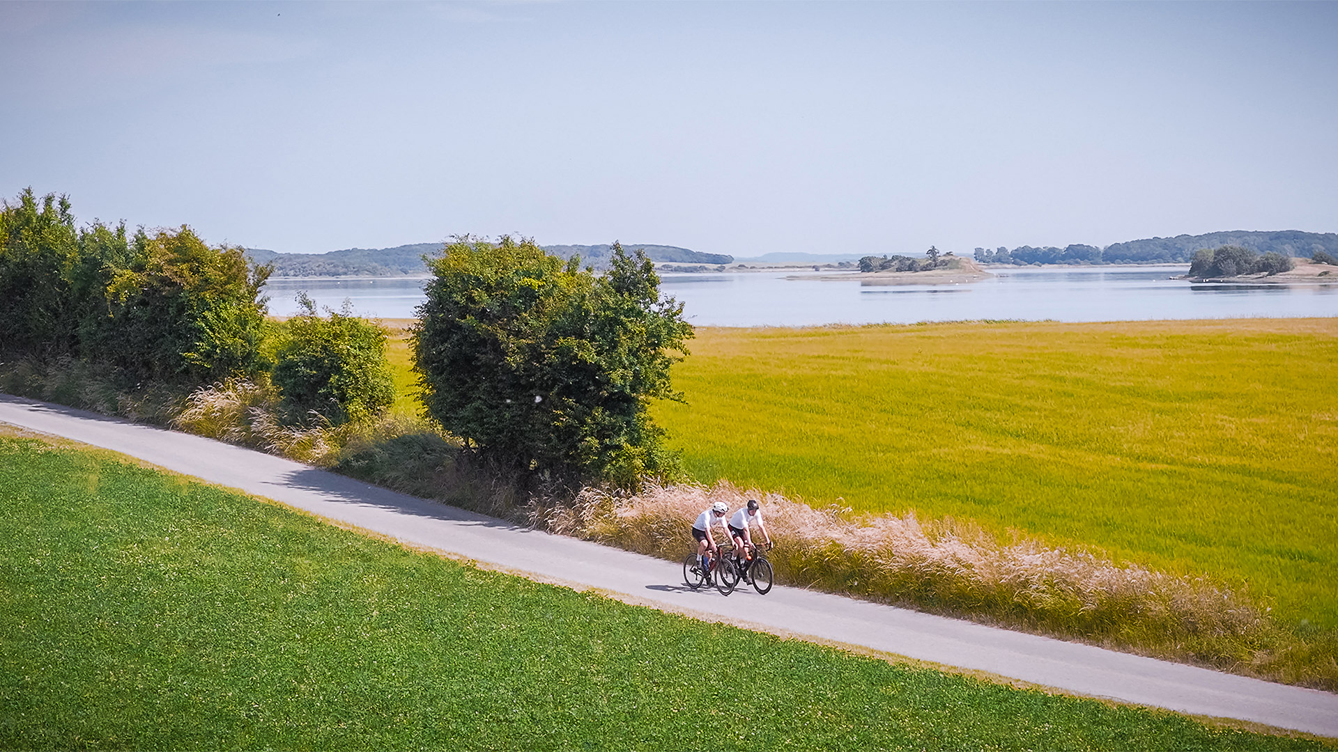 Two cyclists on a country road between green fields with Lindelse Nor in the background