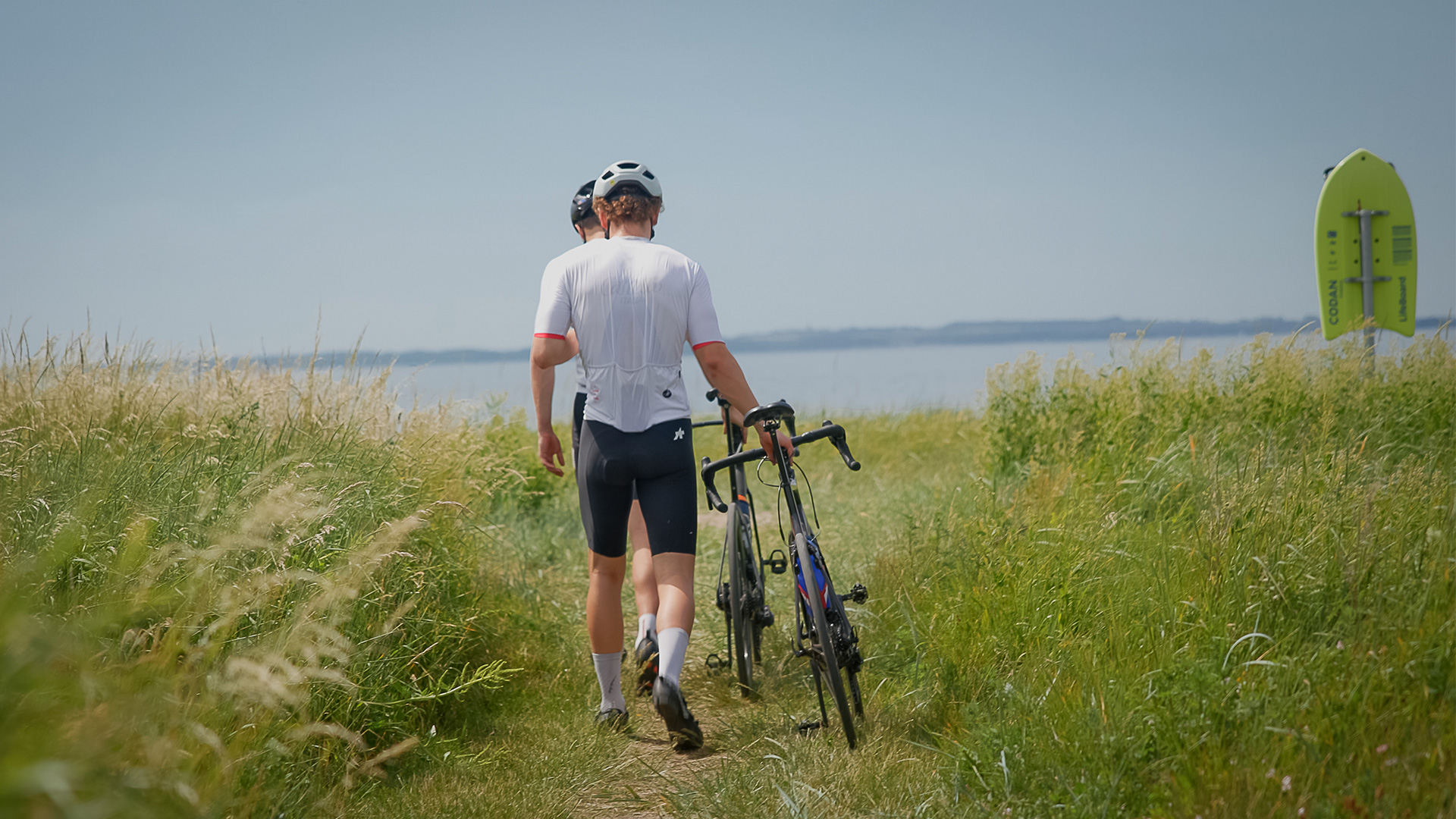 Zwei Radfahrer schieben ihre Fahrräder durch hohes Gras zur Küste auf Langeland
