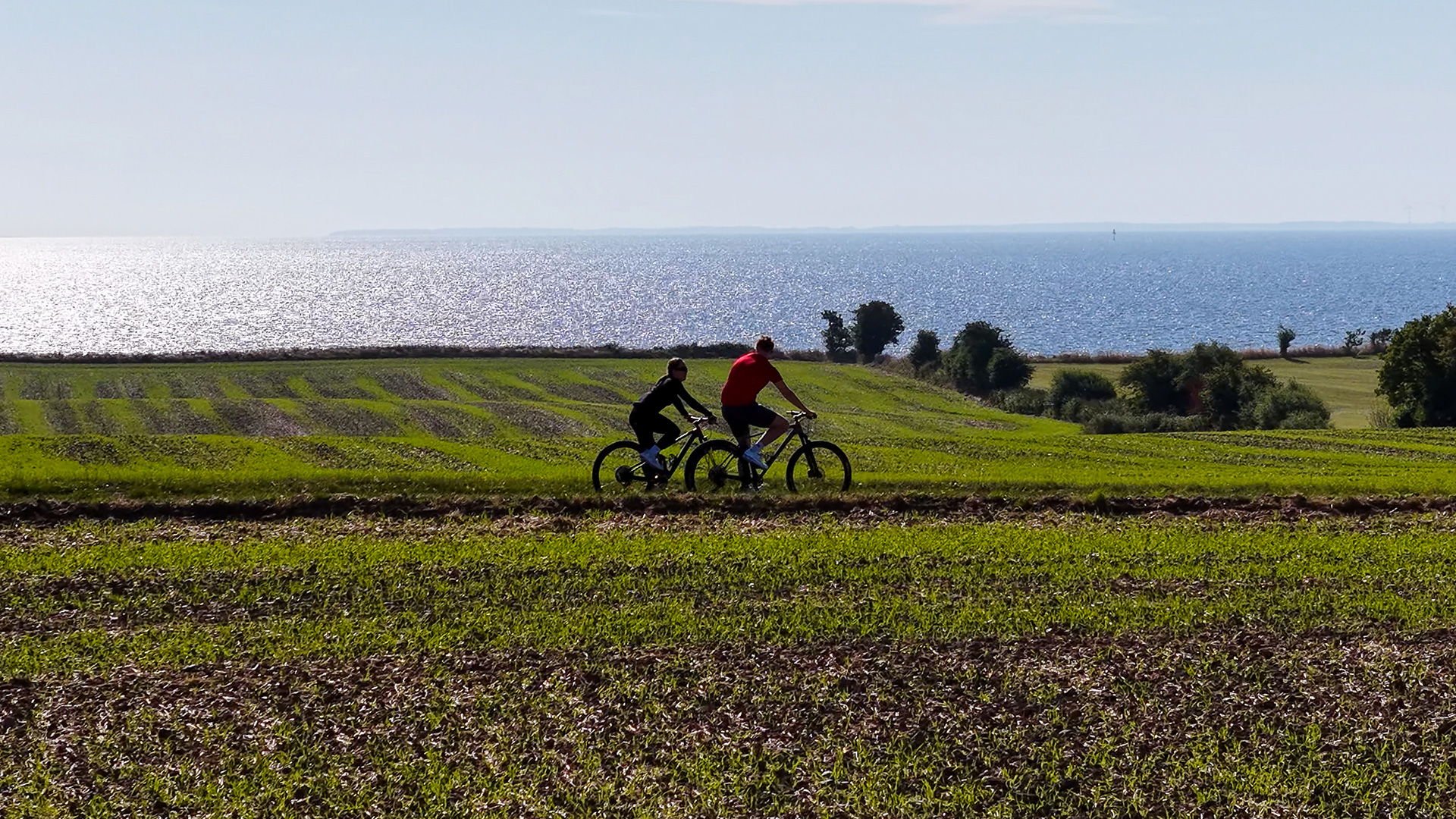 Two cyclists through the open coastal landscape of northern Langeland with the Great Belt in the background