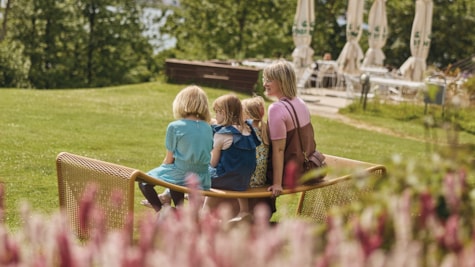 En mor og hendes tre døtre sidder på en træbænk og nyder det gode vejr i Trapholts grønne have.