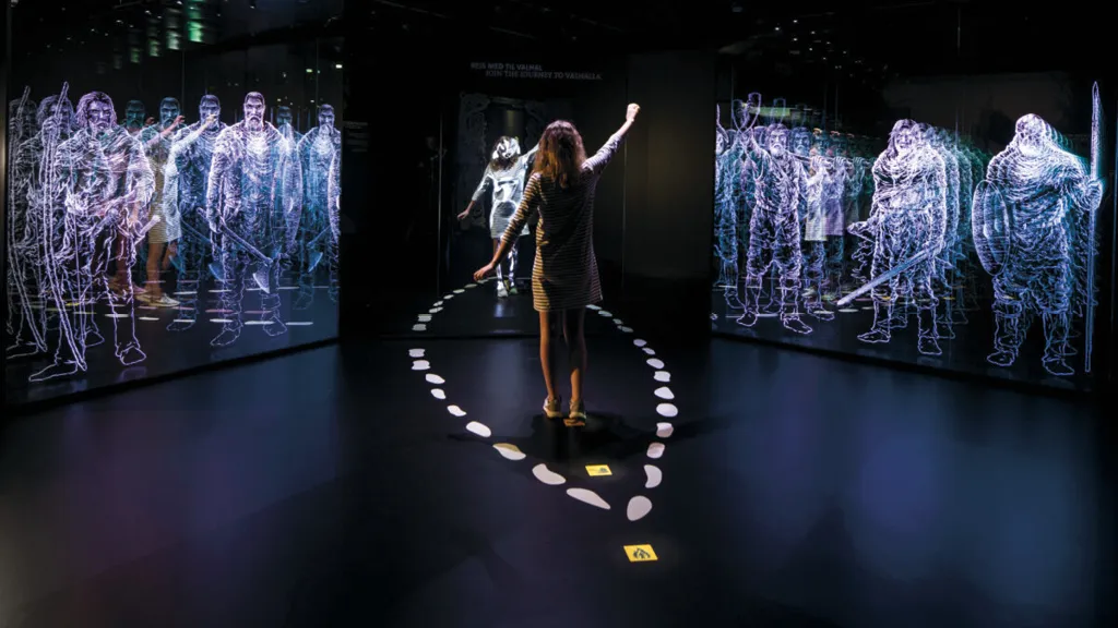 Girl in the interactive exhibition ‘Journey to Valhalla’ at Kongernes Jelling, surrounded by light silhouettes of Vikings.