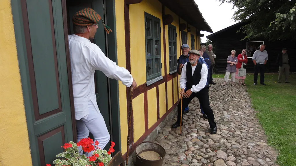 Frivillige optræder i traditionelle kostumer ved gult hus med bindingsværk og stråtag og en brostensbelagt sti til høstfest på Hjortsvang Museum tæt på Uldum og Gudenåen