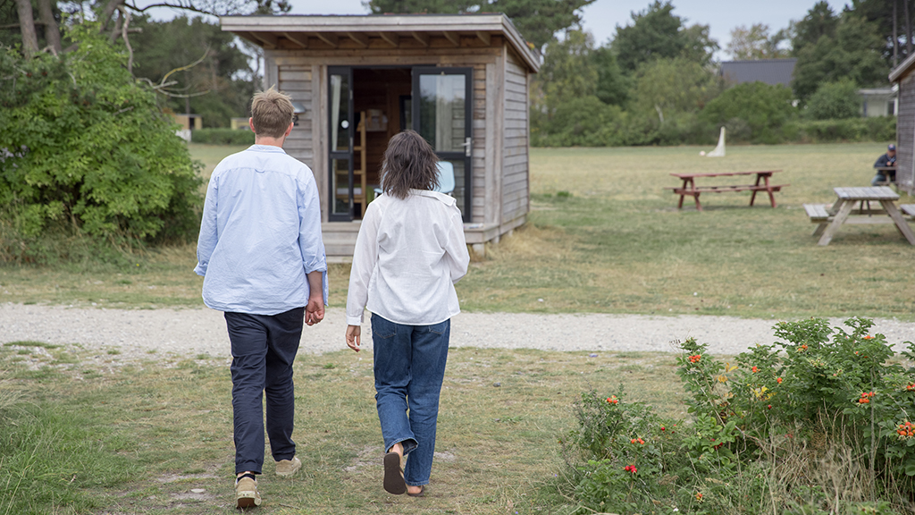 A man and woman are walking towards a cabin on the grass at the Tunø Campsite in Destination Kystlandet