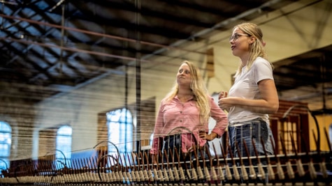 Дві дорослі жінки стоять і дивляться на старий промисловий ткацький верстат у музейній залі Tekstilfabrikken у промисловому музеї в Хорсенсі, Ютландія