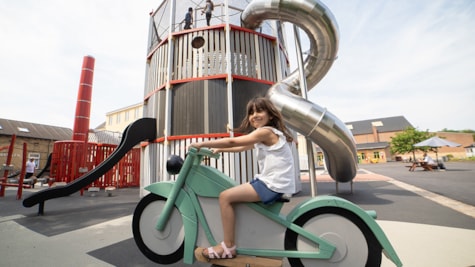 Дитина сидить на зеленому іграшковому мотоциклі на відкритому ігровому майданчику, зробленому Monstrum