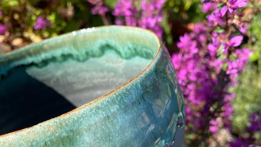 A turquoise-green ceramic coffee cup made by LM Unika in front of a flower hedge