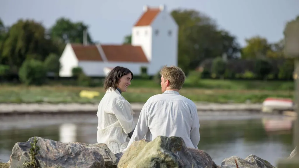 A couple sitting on the beach with Endelave Church in the background