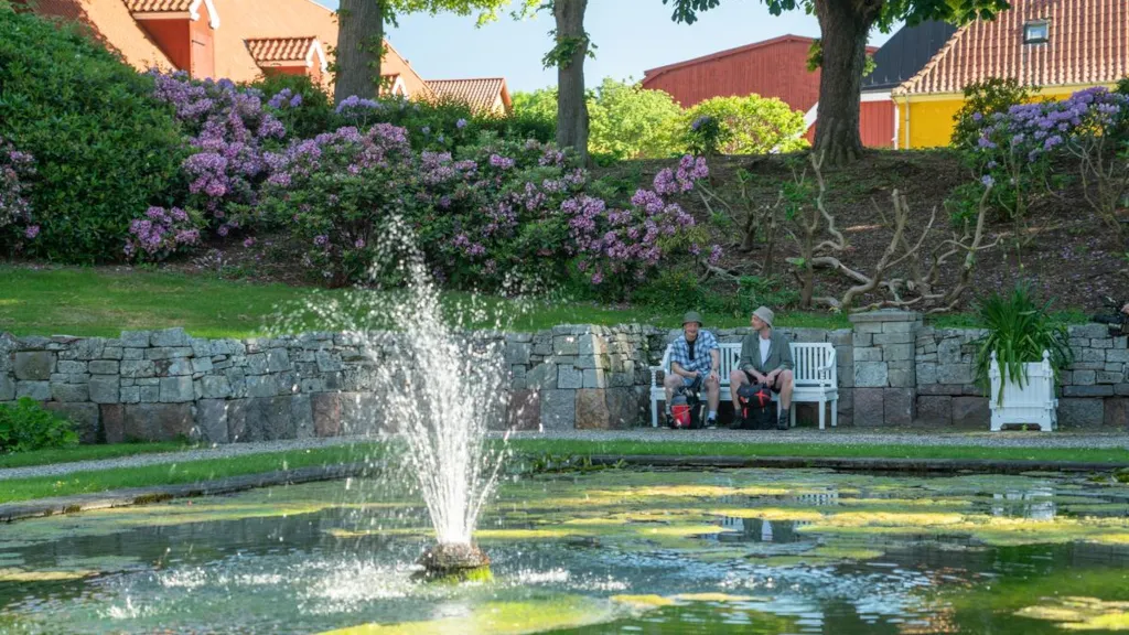 Two men are sitting on a bench in front of a lake with a fountain in Palsgaard Castle Park