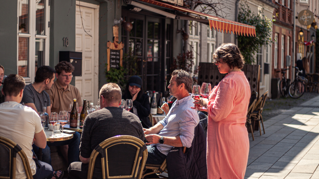 En gruppe sidder i solen ved et bord på Thonbogade foran indgangen til Vores Lille Vinbar
