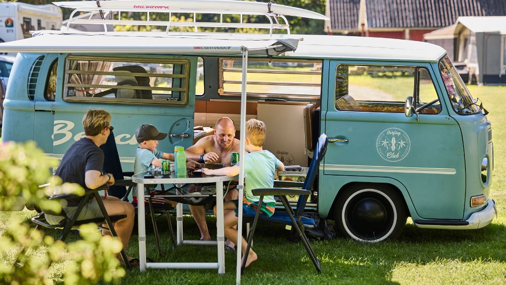 A family sits at the campsite in front of their motorhome – an old VW.