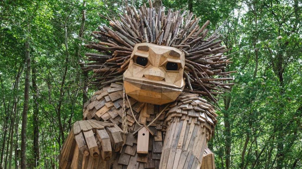 A giant wooden troll with spiky branch hair, built from recycled wood, stands in a green forest