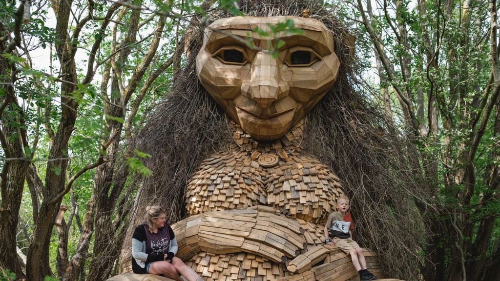 A giant wooden troll with long twig hair sits in the forest, with two people resting on its arms