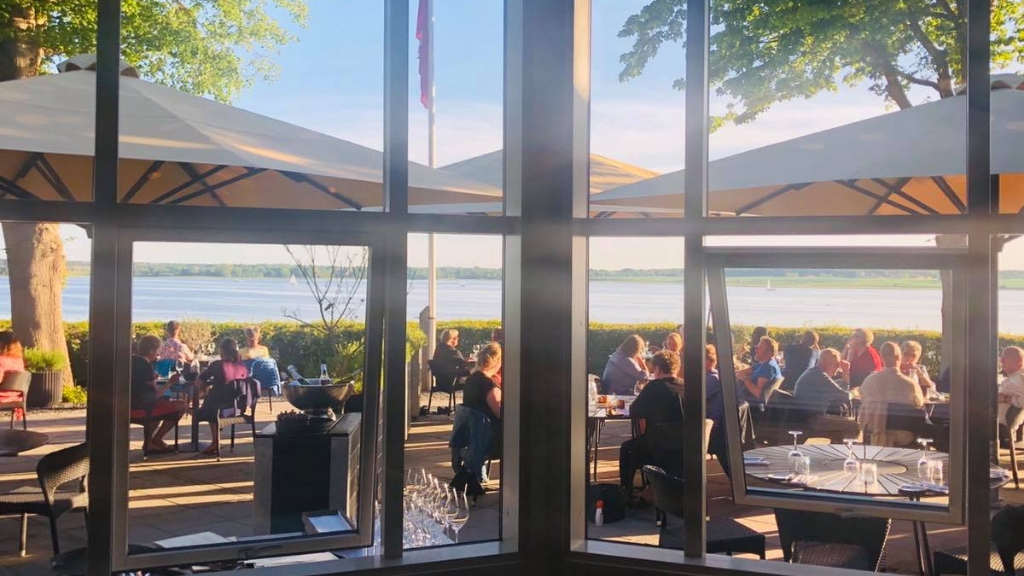 Gæster under parasoller på Restaurant Vigens terrasse ved Roskilde Fjord, set gennem vinduerne.