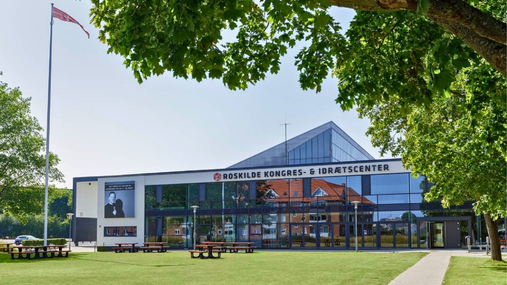 Facade of Roskilde Kongres- og Idrætscenter with green areas and picnic tables.